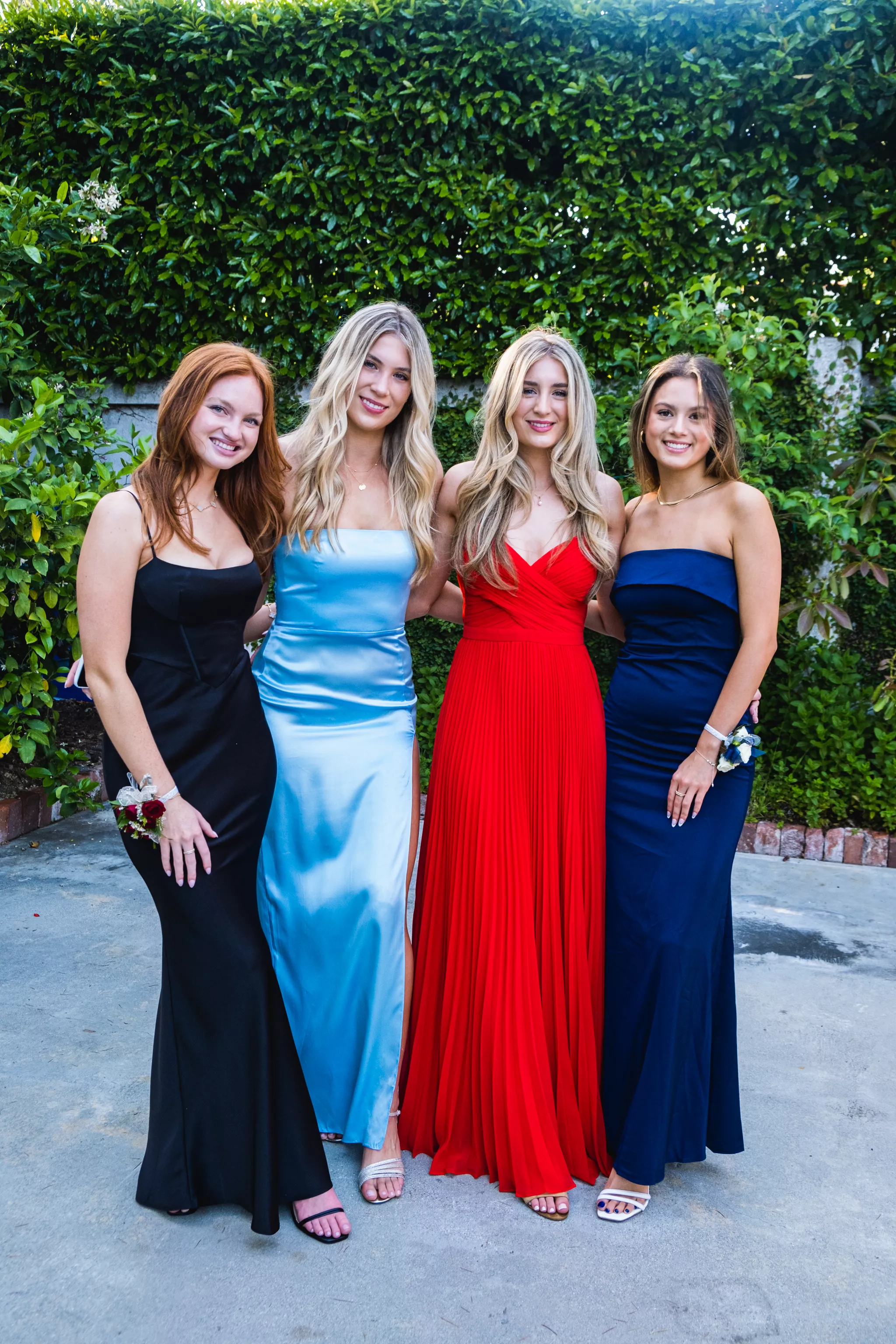 Prom poses for a group of friends — four in formal gowns against a hedge in soft shade