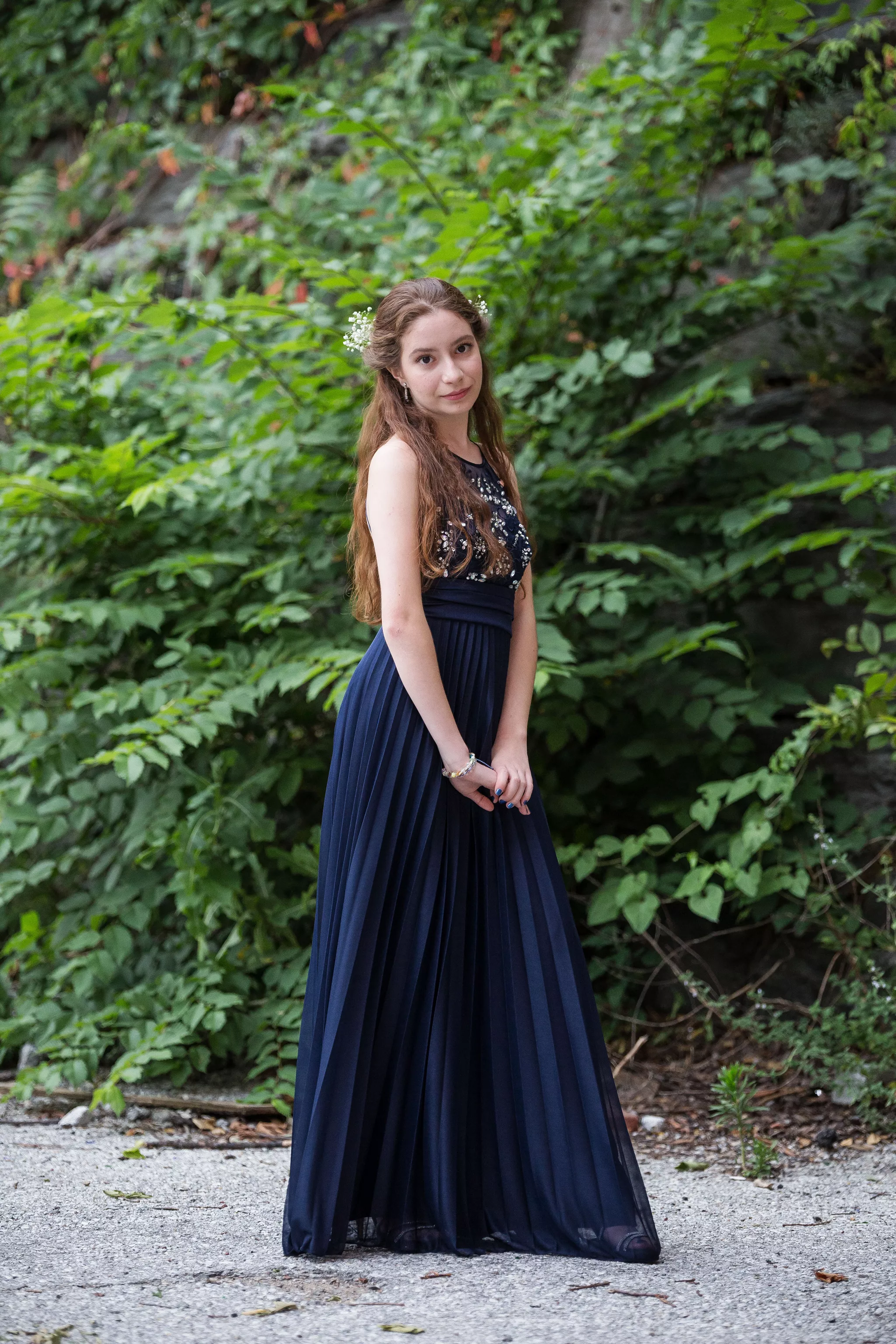 Prom poses in a navy gown — body turned side-on, face back to the camera by an ivy wall