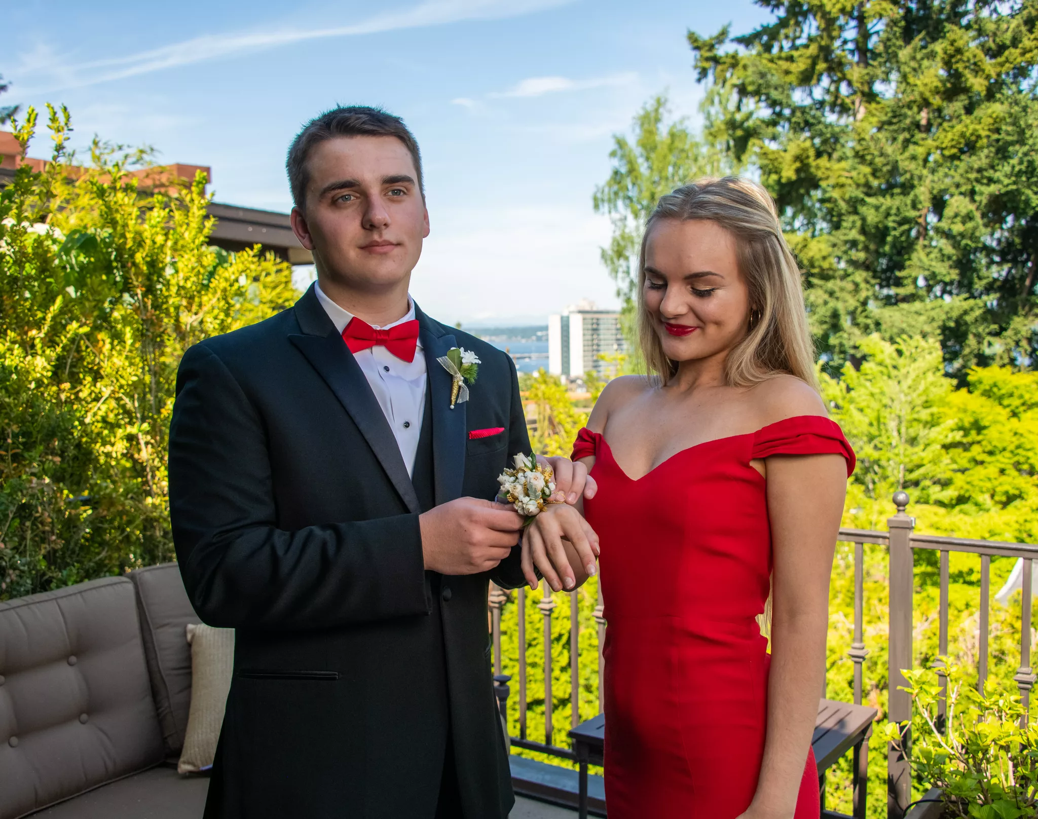 Prom poses with a corsage — he slips a white rose corsage onto her wrist on a sunny deck