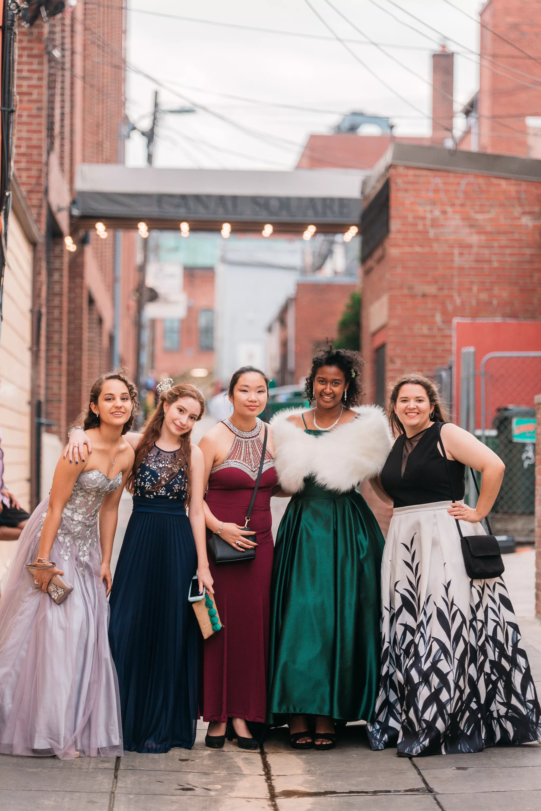 Group prom poses — five friends in gowns arm in arm in a brick alley under string lights