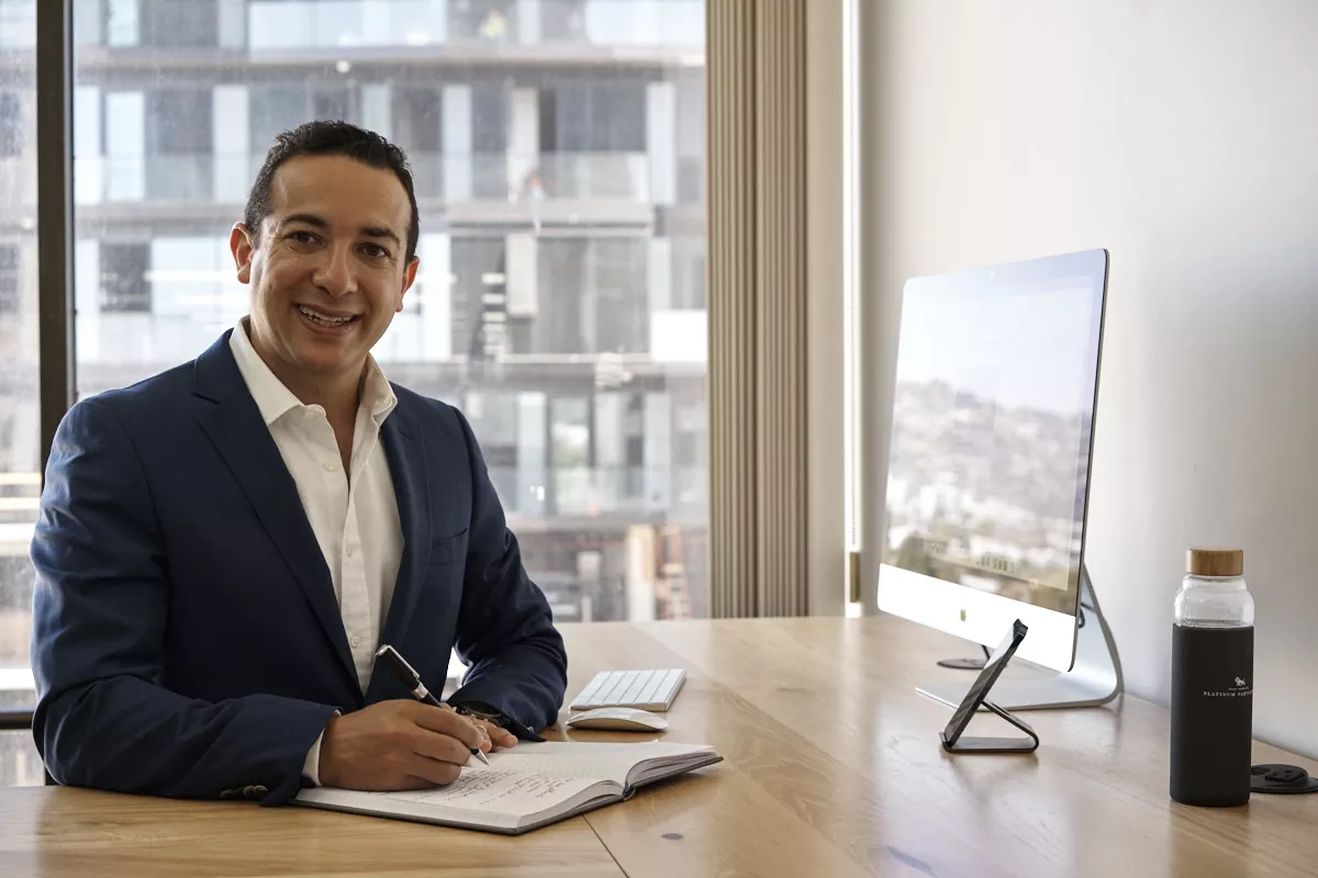 Headshot poses at a desk — navy blazer, body turned off-square, face back to the camera.