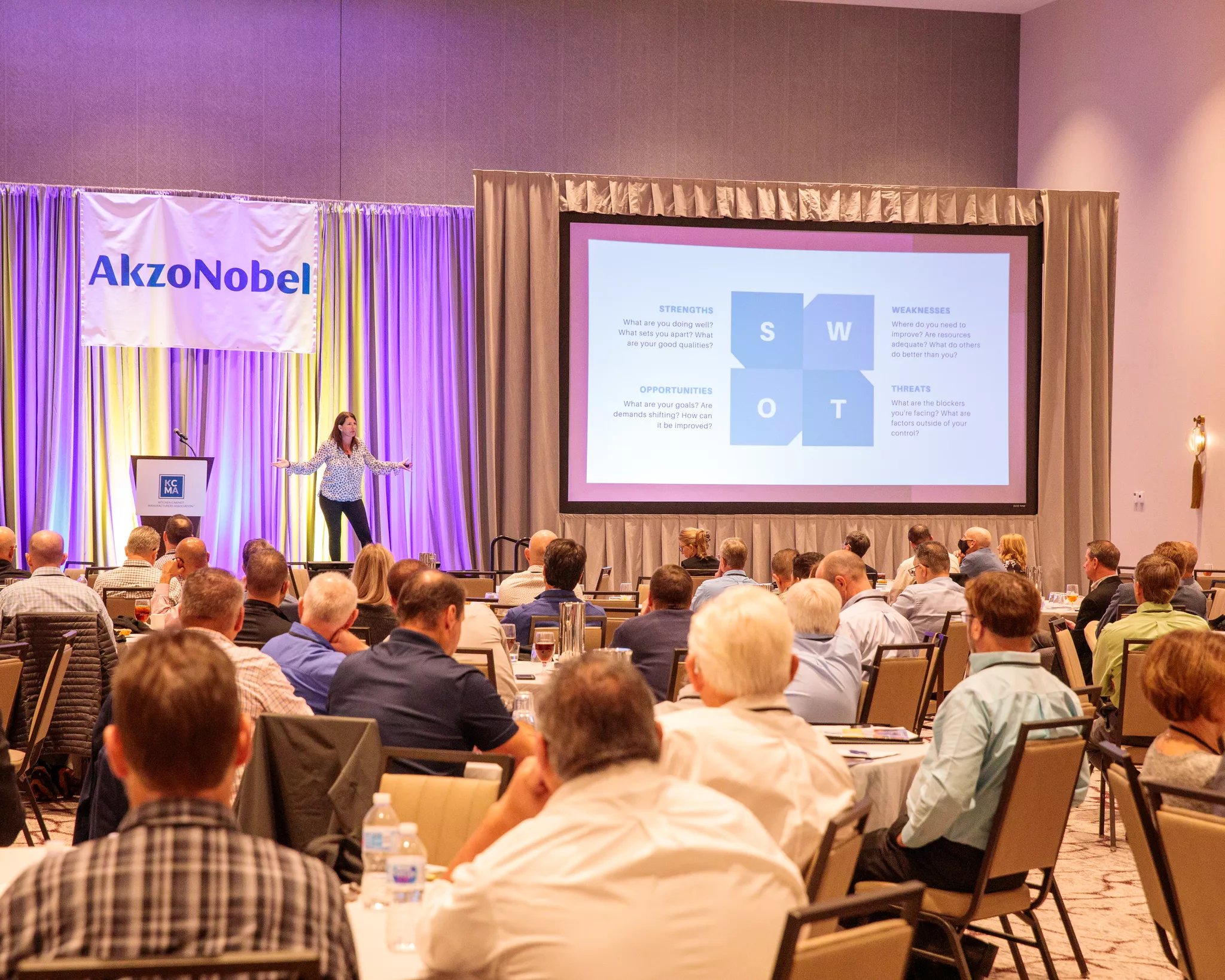 Conference photography of a speaker presenting a SWOT slide beside an AkzoNobel sponsor banner