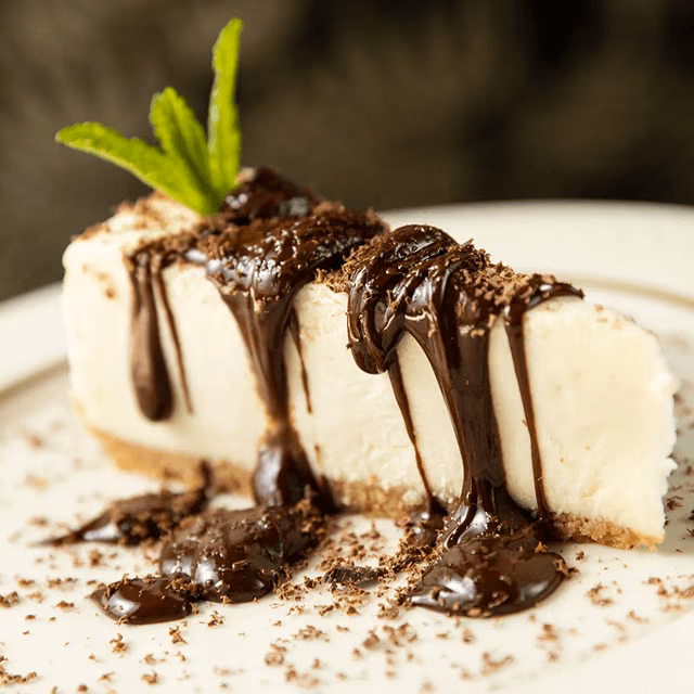Slice of vanilla cheesecake topped with melted chocolate sauce and fresh mint