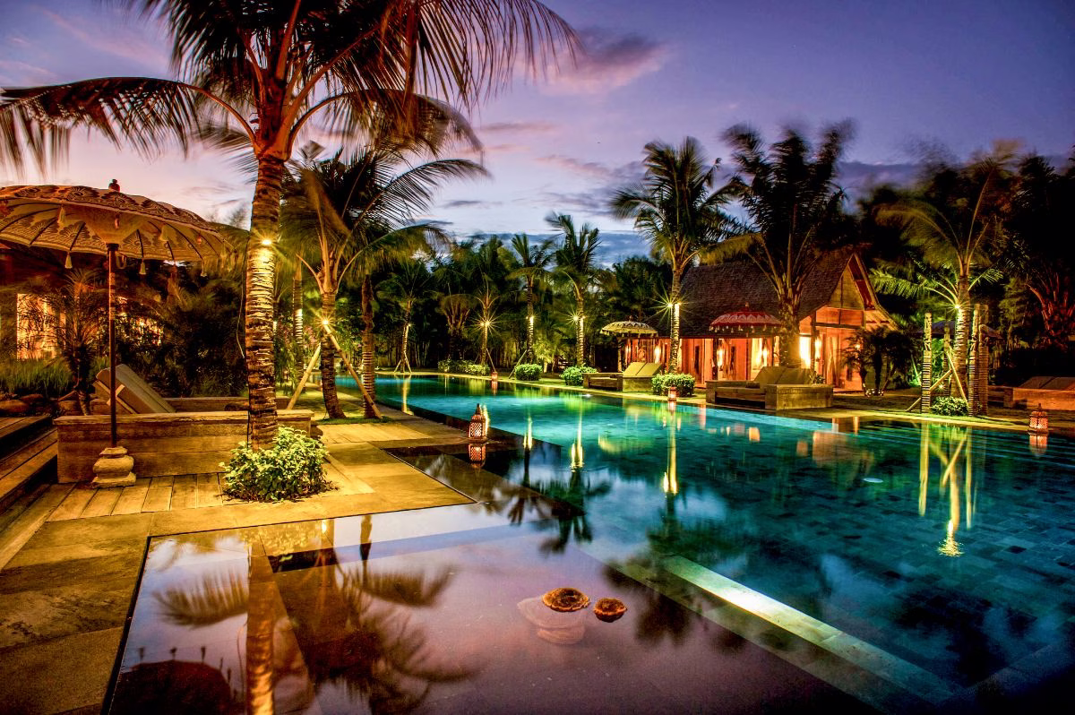 Professional real estate photo of a tropical resort pool at dusk lined with palm trees and lanterns