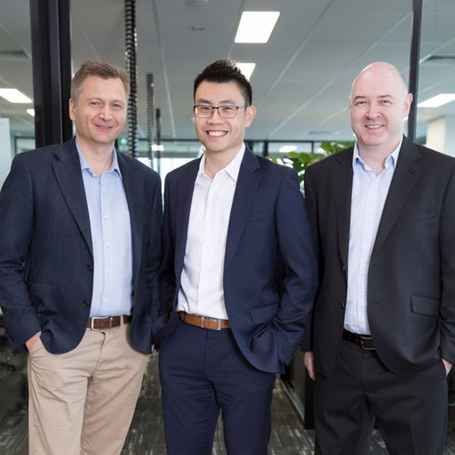 Three businessmen in suits smiling together in a professional portrait inside a modern office.