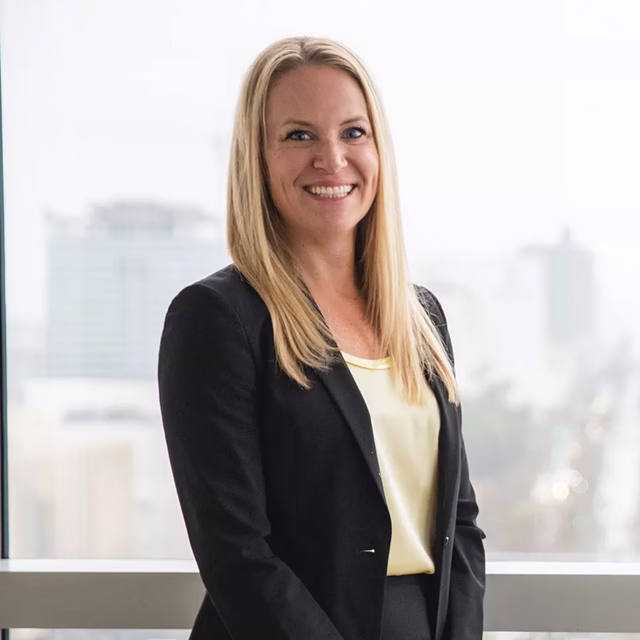 Blonde woman in a black blazer posing for a professional headshot next to a city-view window.