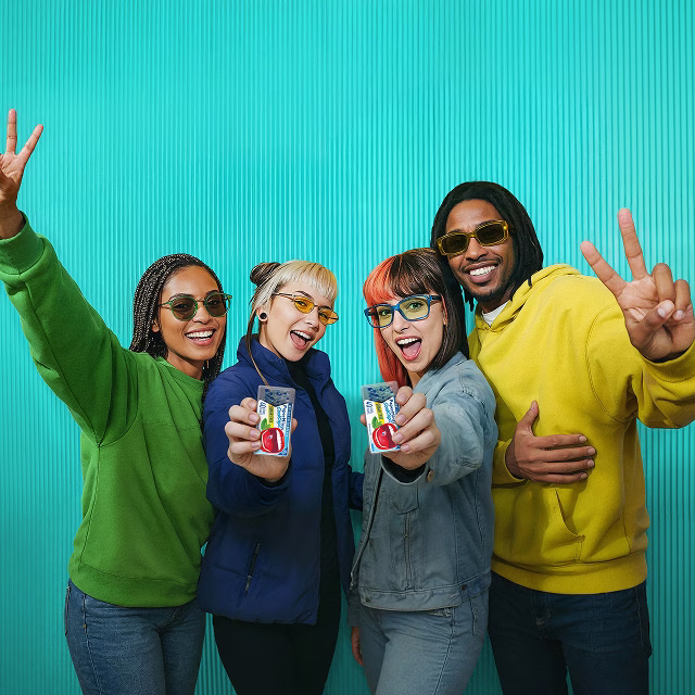 Four friends in colorful hoodies holding a product and smiling against a teal background.