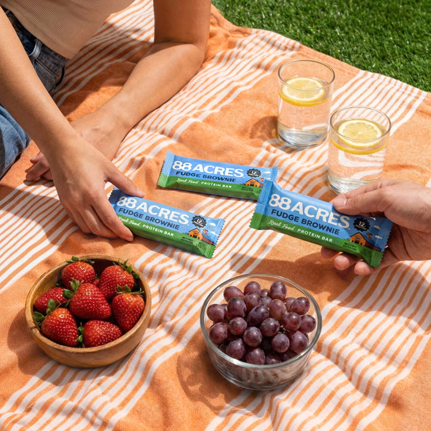 Two people sharing a protein bar on a picnic blanket surrounded by strawberries and grapes.