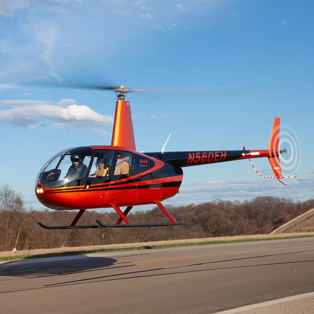 Red Robinson R44 helicopter lifting off from a road with a pilot at the controls.