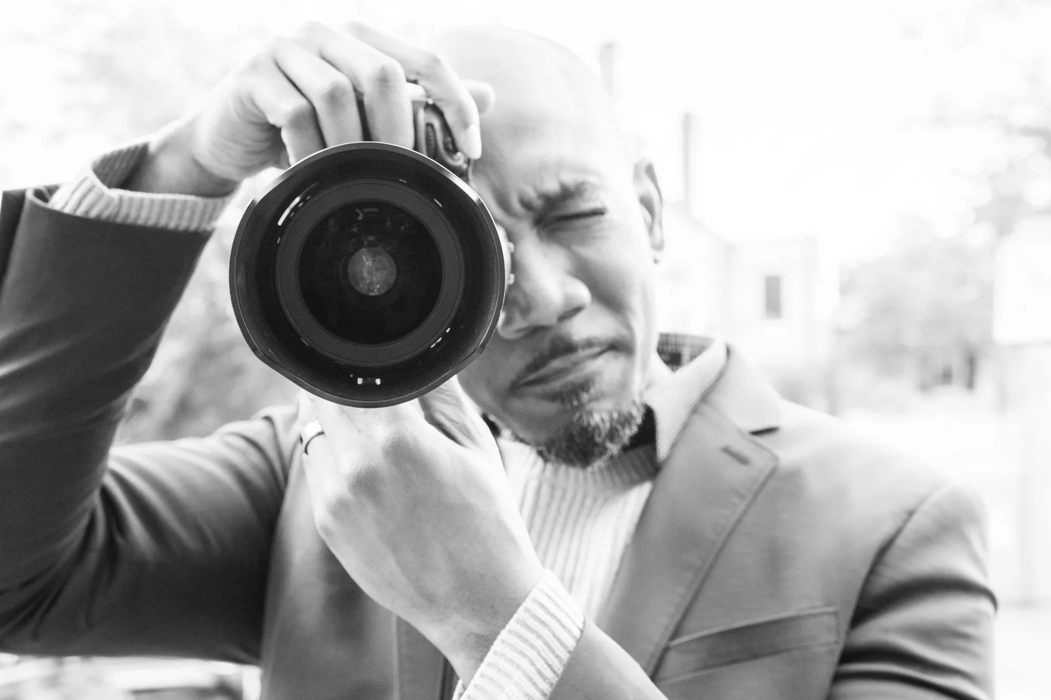 Photographer in a blazer aiming a DSLR lens straight at the camera, black and white