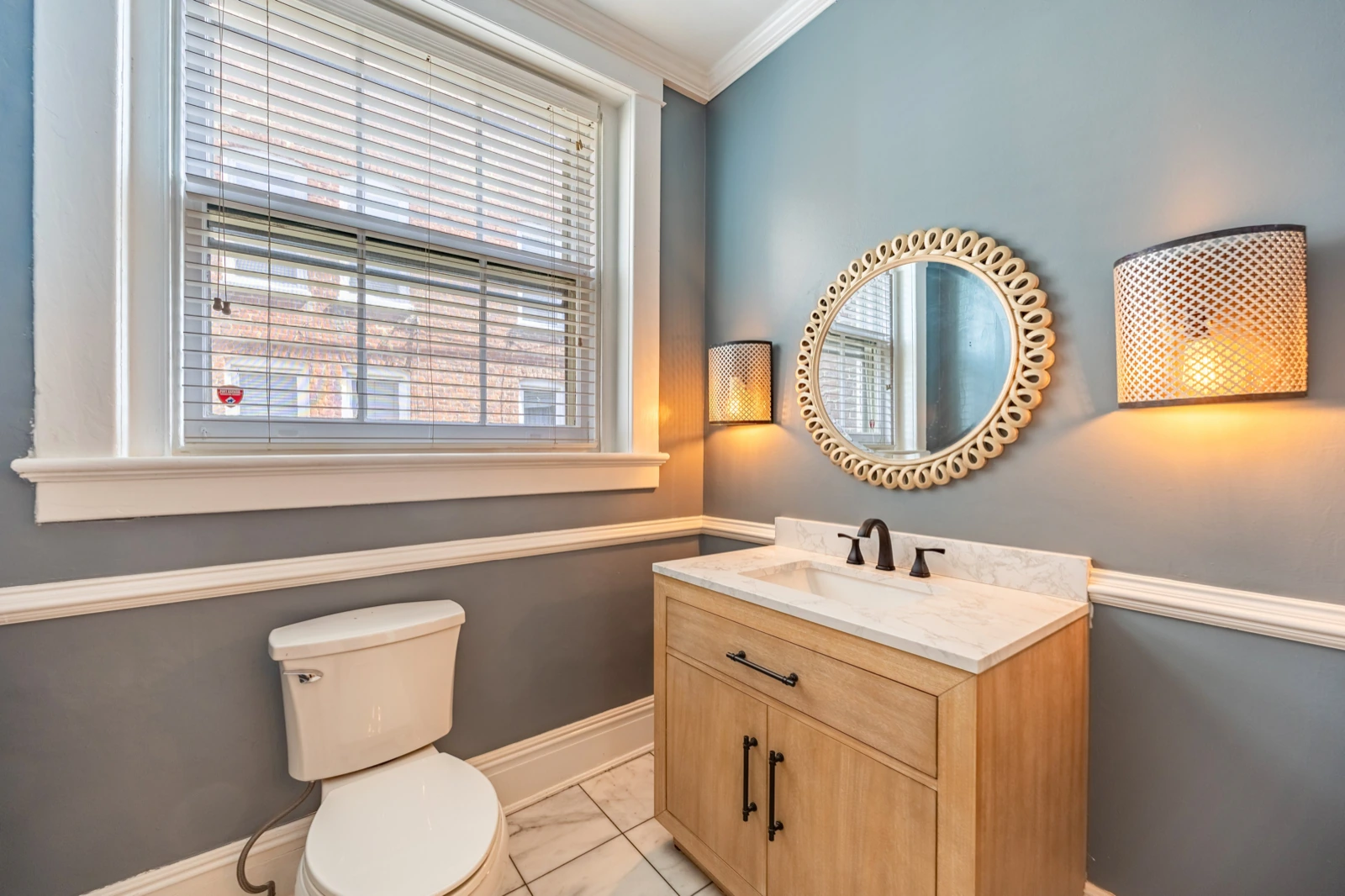 Bathroom with a wood vanity, round mirror and lit wall sconces, toilet lid closed