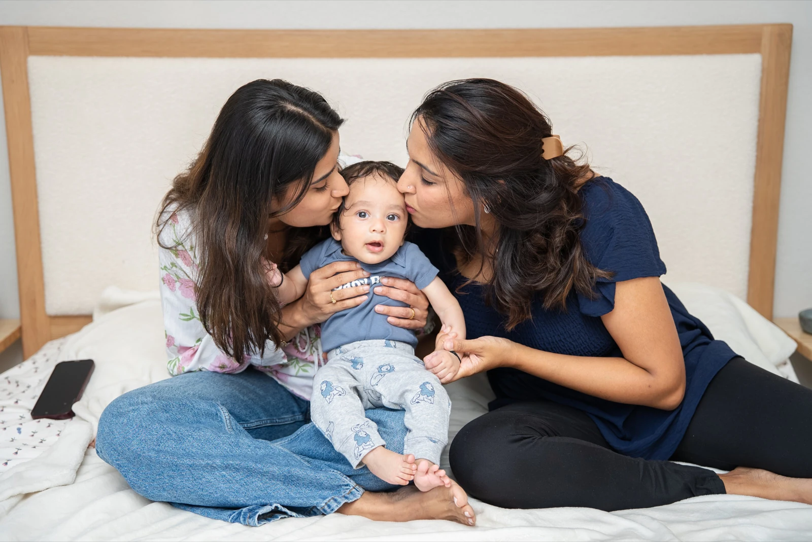 Family photo poses with a baby: two parents kissing their seated baby on a bed