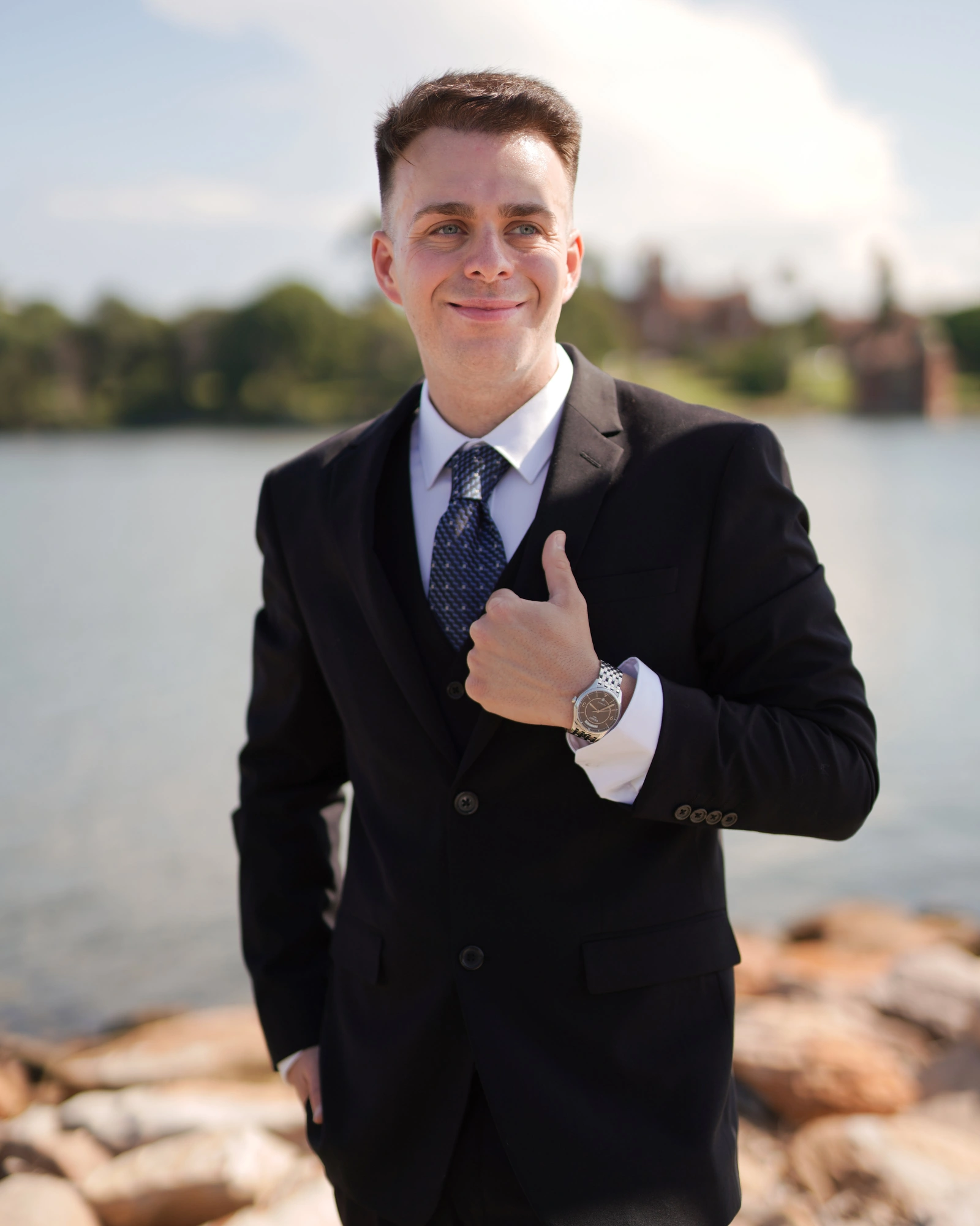 Male poses with layers: man in a dark suit holding his jacket lapel by a lake