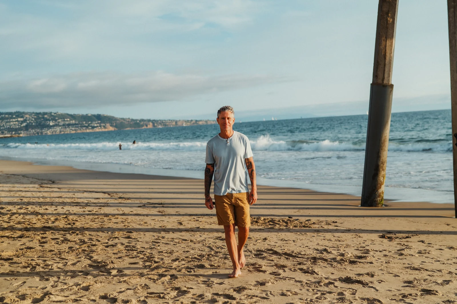 Outdoor male poses: man walking barefoot toward camera on a sandy beach by a pier