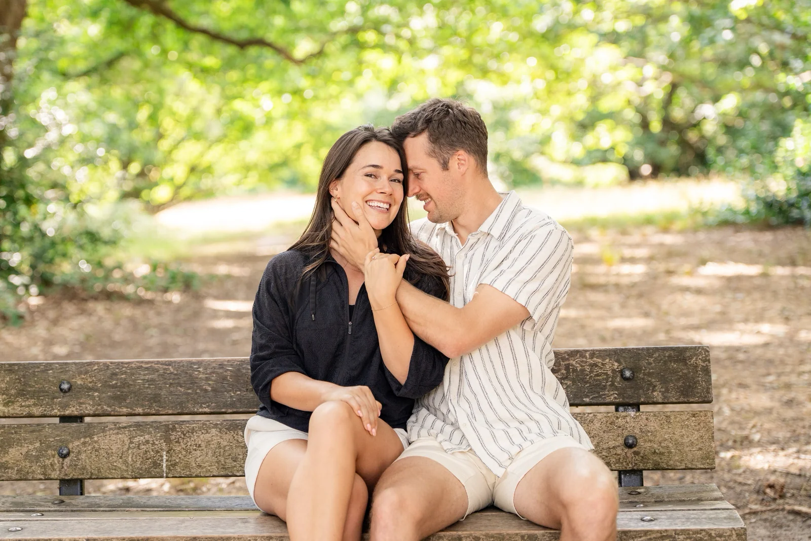 Candid couple poses on a park bench, one partner laughing at a whispered prompt