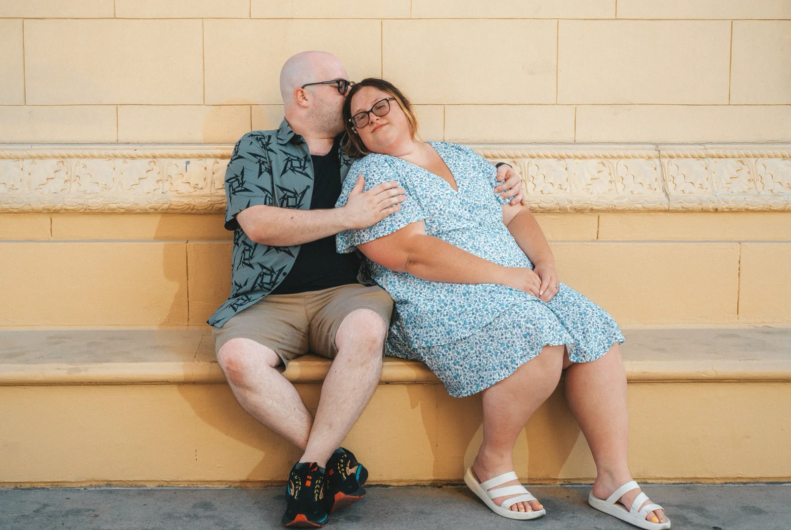 Seated couple poses on a stone ledge, with a kiss on the top of the head