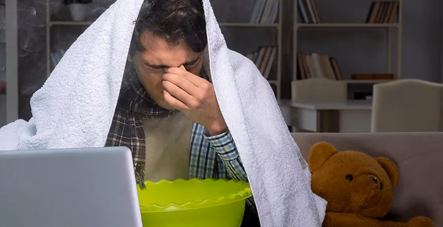 Person with towel over head above steam bowl, laptop in front, teddy bear on couch.