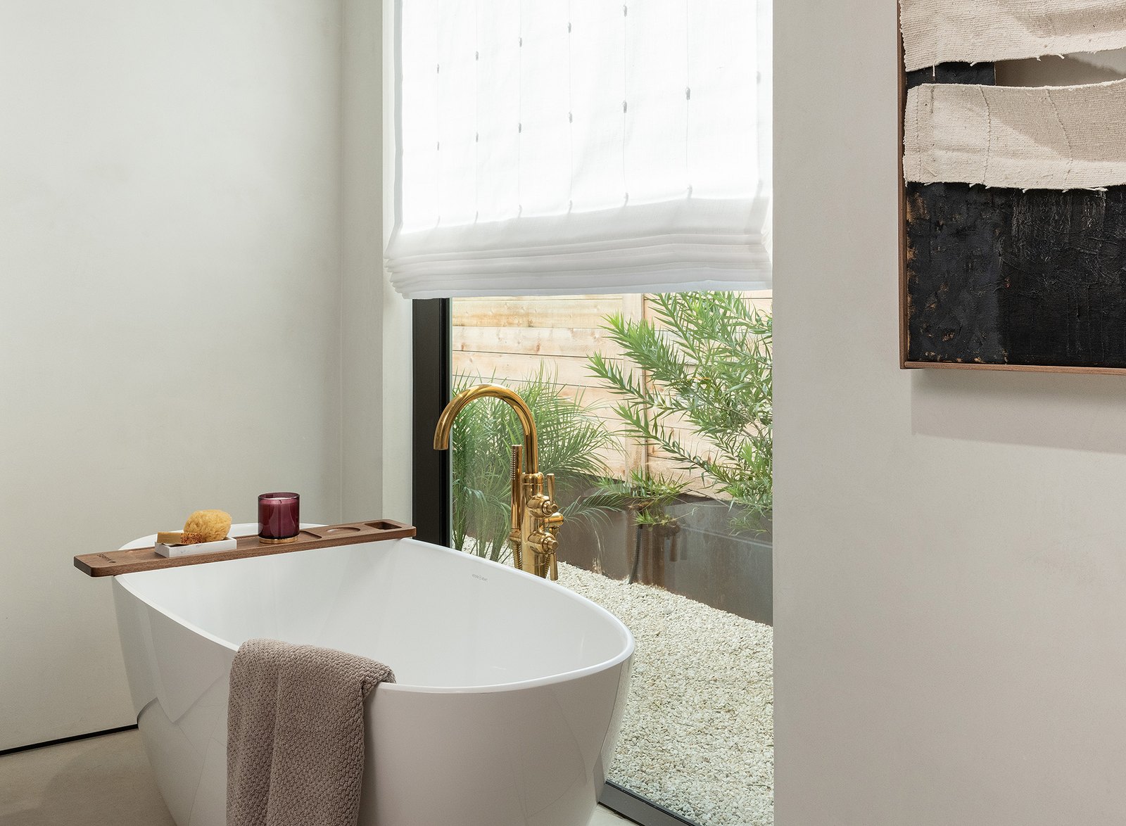 Bathroom with a white Roman shade above the window, freestanding tub, brass floor mounted filler, gravel courtyard beyond