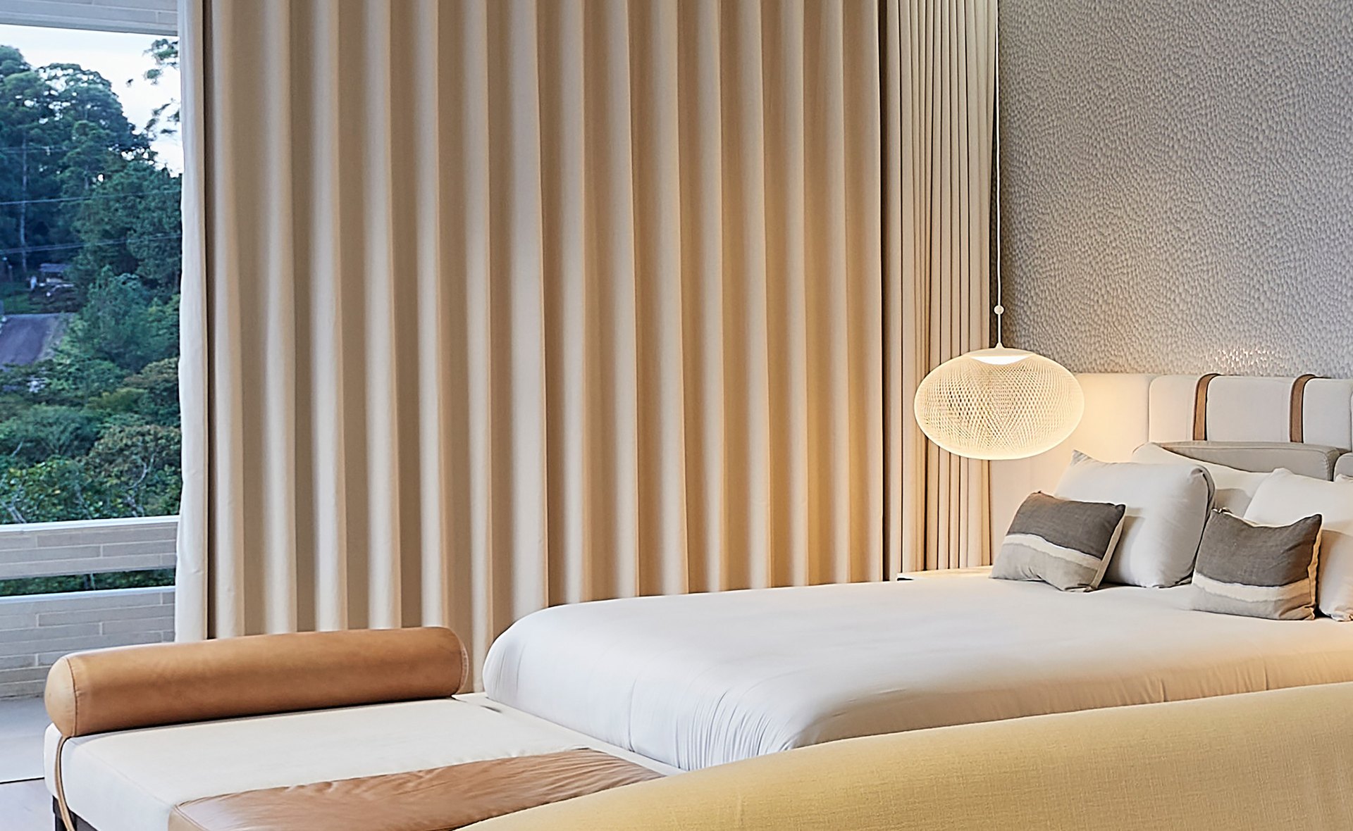 Bedroom with full height cream drapery, bed with bolster bench, woven globe pendant, treetops through the glass