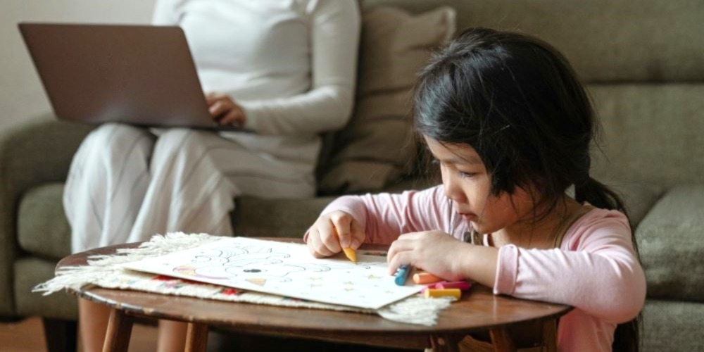 A little girl is drawing at a coffee table while a woman sits behind her with a laptop.