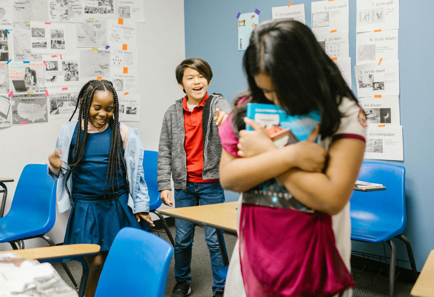 In a classroom, a girl holds her book self-consciously while two other students stare at her.