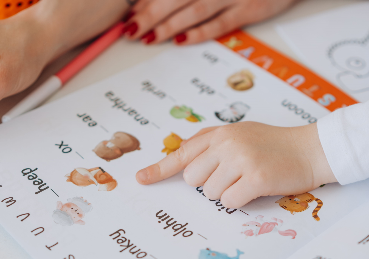A child's hand points at a worksheet of words including animal names