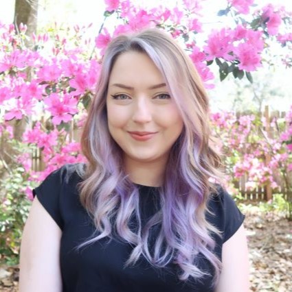 A headshot of Kristen smiling. She has brown and purple curly hair and is standing in front of a bright pink azalea bush.