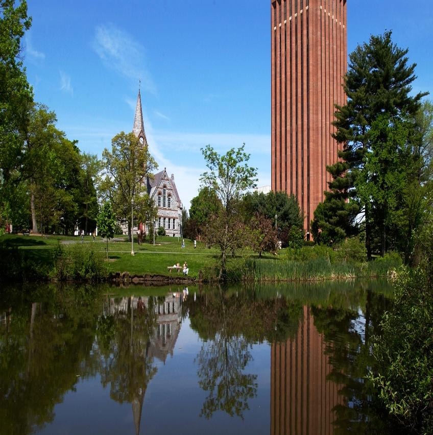 The image depicts a serene park scene with a reflective pond, tall red brick church steeple, and surrounding trees.AI-generated content may be incorrect.