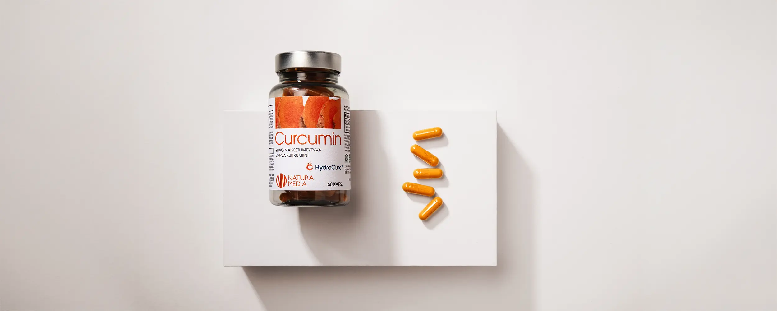 Natura Media Curcumin -purkki on aseteltuna kyljelleen vaalean beigelle korokkeelle. Korokkeella on myös aseteltuna 5 kirkkaan oranssia Curcumin-kapselia.