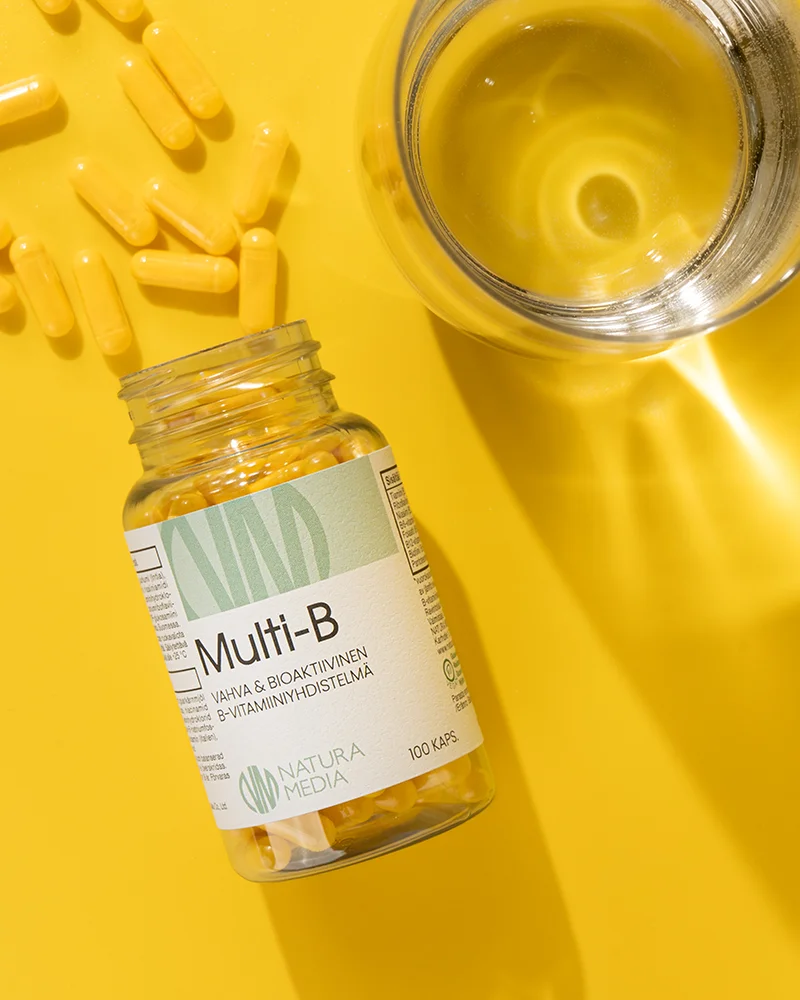 Avattu Multi-B purkki makaa keltaisella pöydällä, jossa on aseteltuna myös keltaisia Multi-B-kapseleita ja lasillinen vettä.