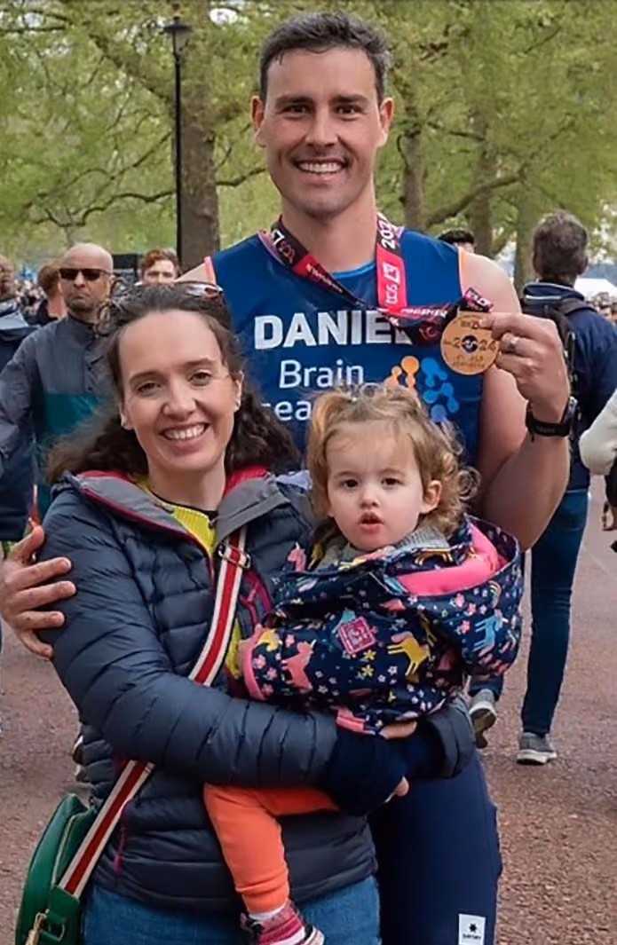 Becky with their youngest daughter on her arm, standing beside Dan who's holding a London Marathon medal