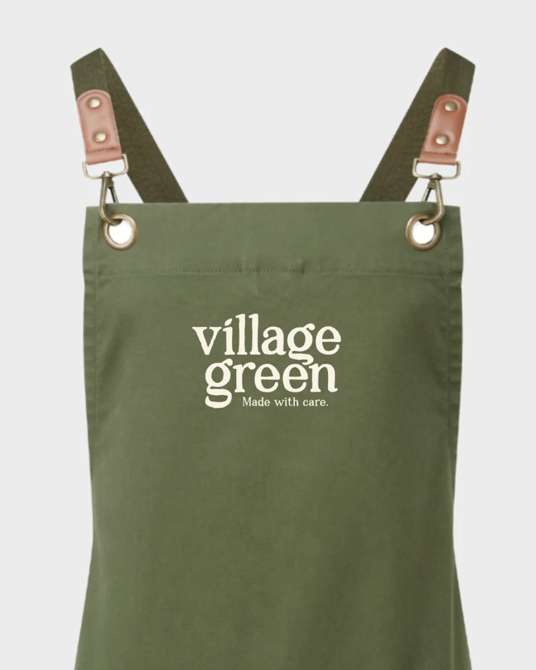 Green apron with brown straps and white text reading 'village green Made with care.' on front.
