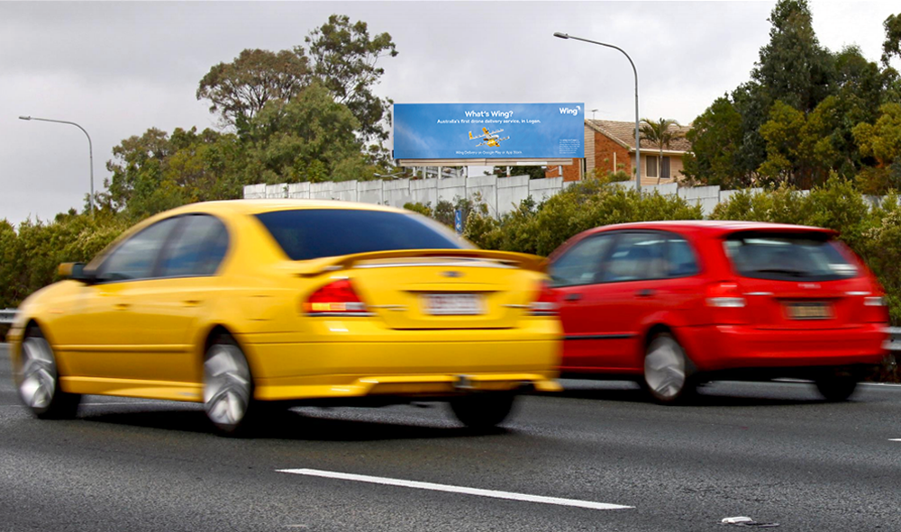 Image of a billboard on-site above a busy highway. The billboard featuring photography of a Wing commercial drone. The billboard says "What's Wing? Australia's first drone service, in Logan. Wing Delivery on Google Play or App Store."