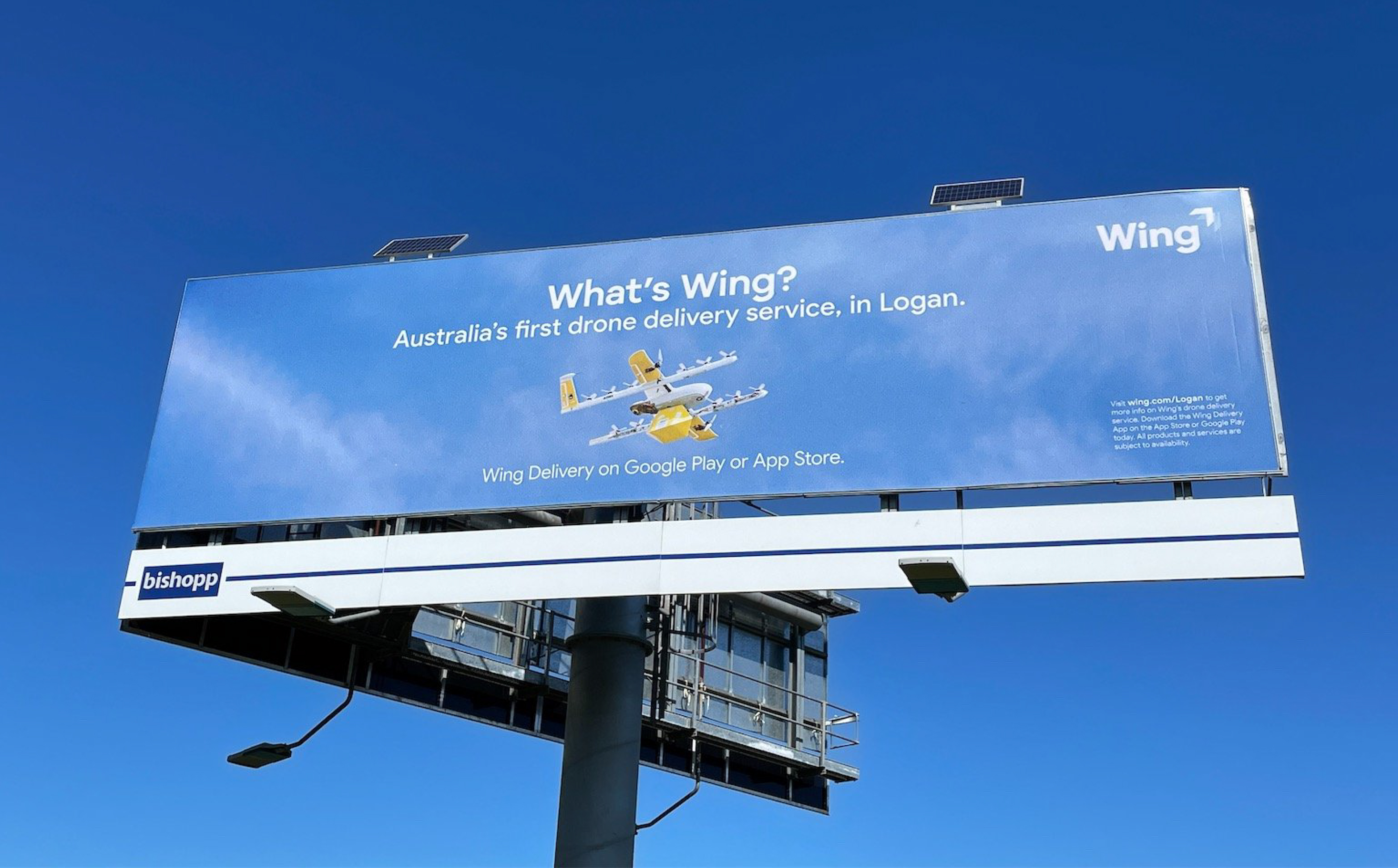 Image of a billboard on-site in a clear blue sky. The billboard featuring photography of a Wing commercial drone. The billboard says "What's Wing? Australia's first drone service, in Logan. Wing Delivery on Google Play or App Store."