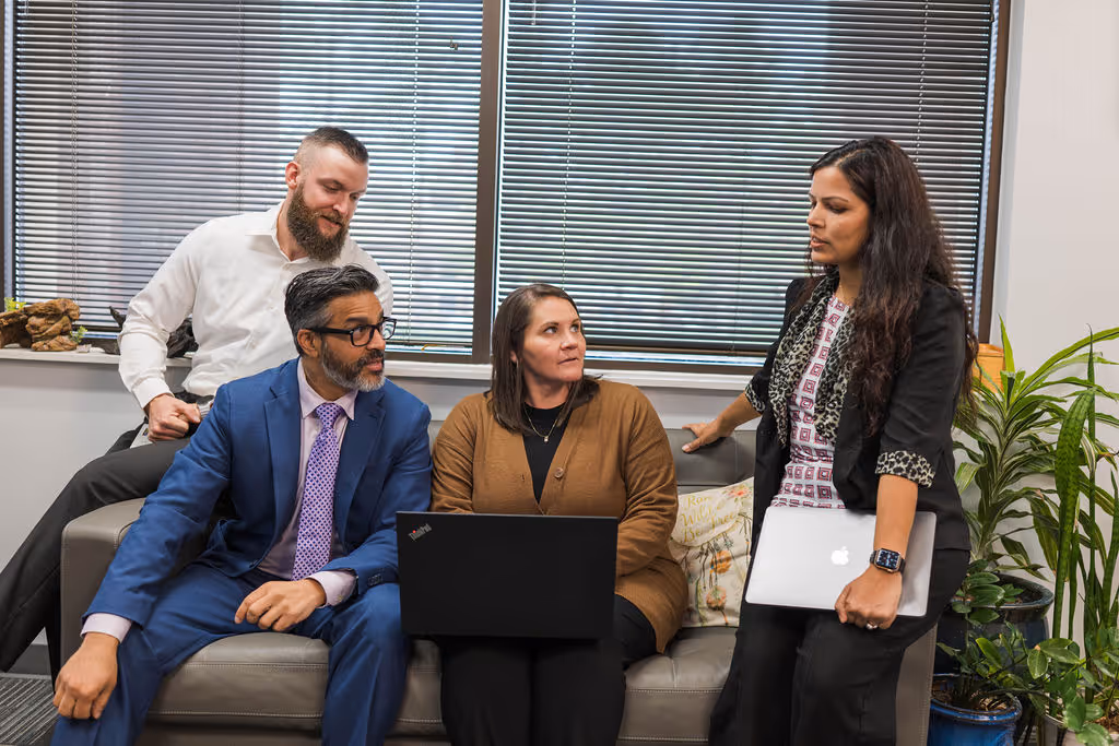 Four diverse colleagues having a discussion in an office, three seated on a couch with laptops and one standing nearby.