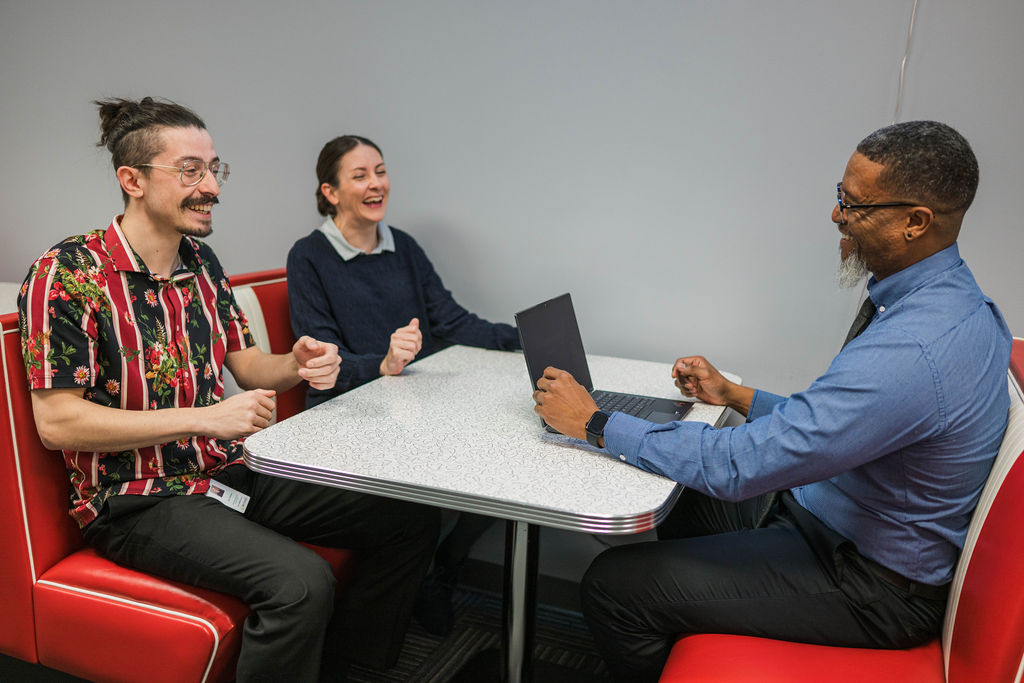 Three colleagues smiling and laughing while sitting around a table, with one using a laptop.