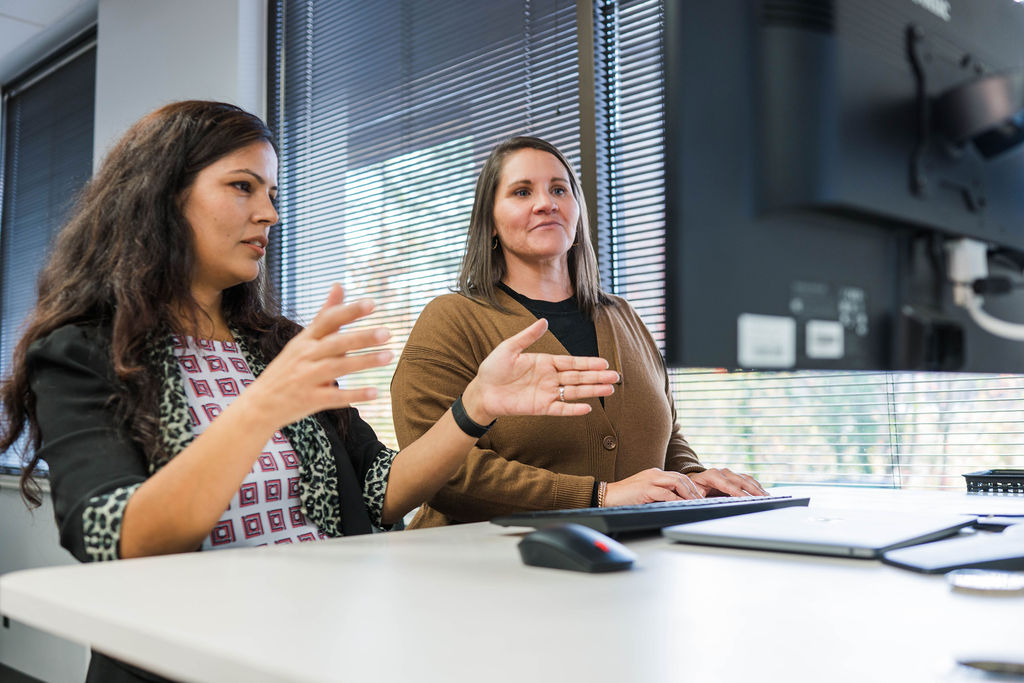 Two women at a desk discussing work in an office with a computer monitor and keyboard.