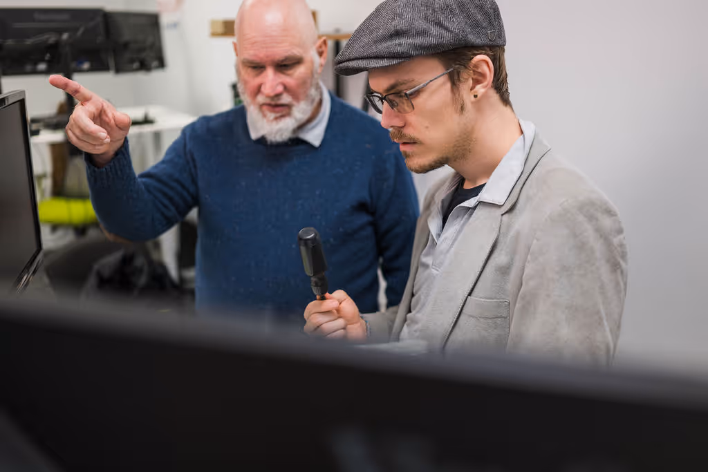 Older man in blue sweater pointing at a computer screen while younger man with glasses and a flat cap holds a screwdriver and looks focused.