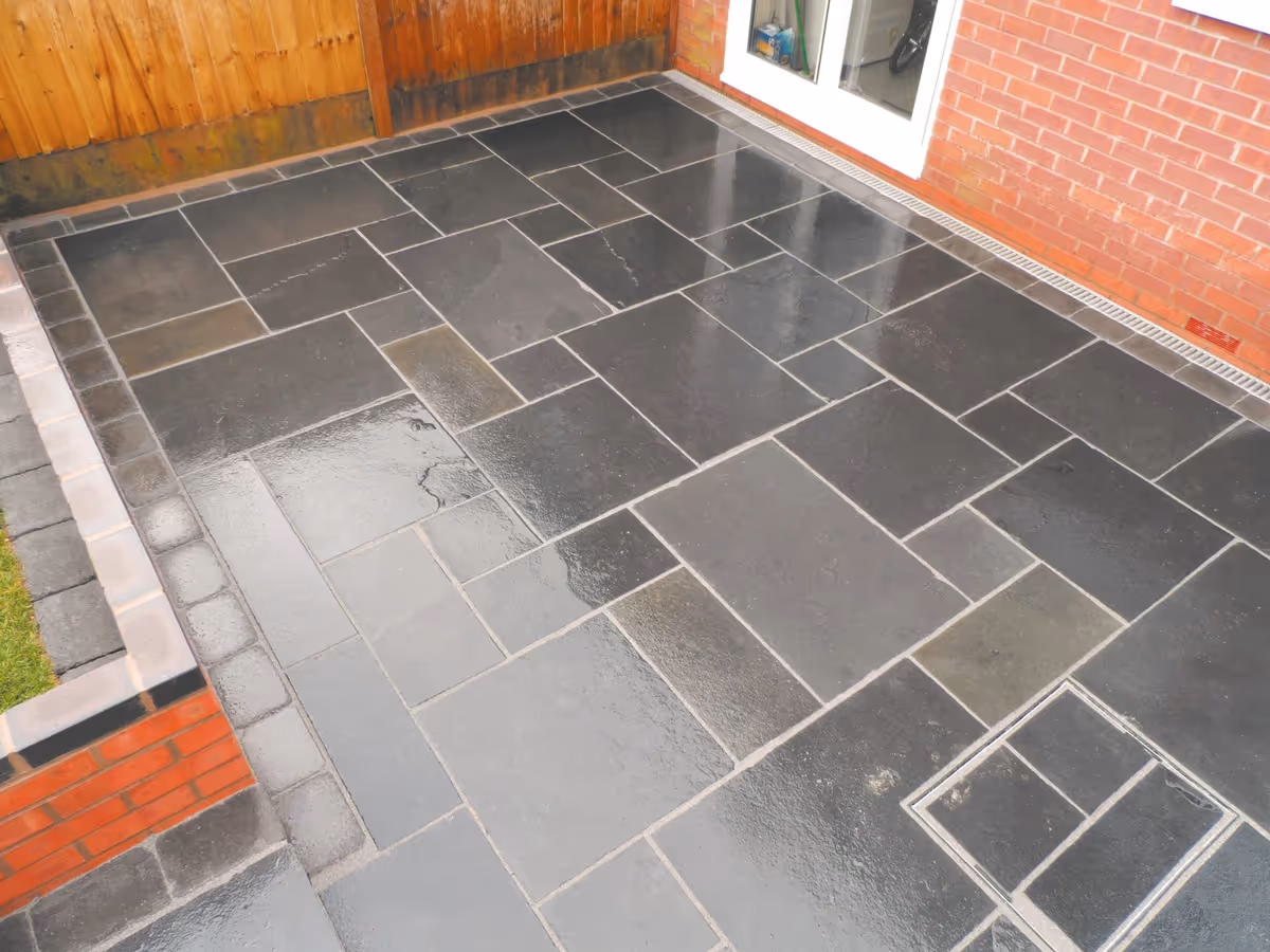 Mixed size black limestone patio with large charcoal rumbled block, red walling finished with blue engineering brick, new lawn and treated feather edge fencing - angle 1