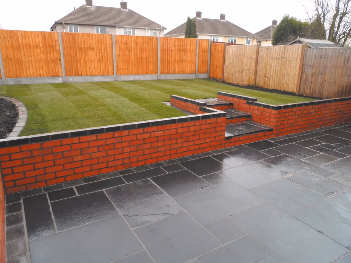 Mixed size black limestone patio with large charcoal rumbled block, red walling finished with blue engineering brick, new lawn and treated feather edge fencing - angle 3