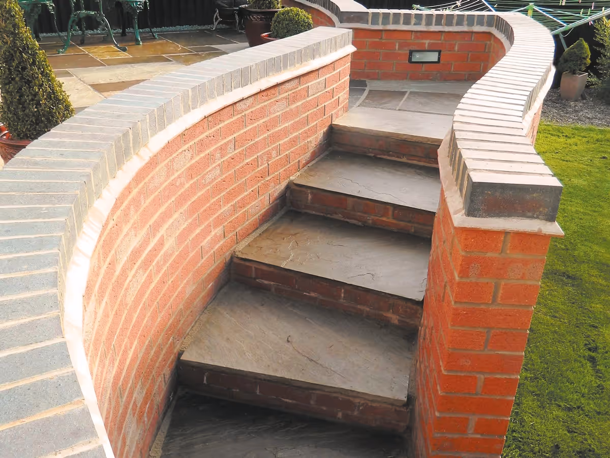 Mixed size camal natural stone patio with large charcoal rumbled block, large red retaining wall with blue engineering brick - angle 2