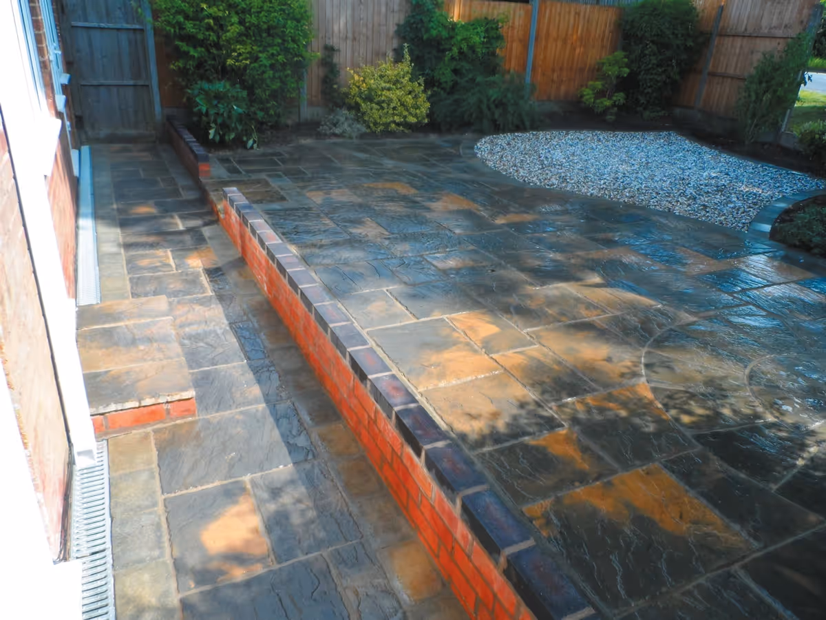 Winter stone concrete patio with matching circle, patio edged with large charcoal rumbled block, red walling finished with blue engineering brick and new lawn - angle 2