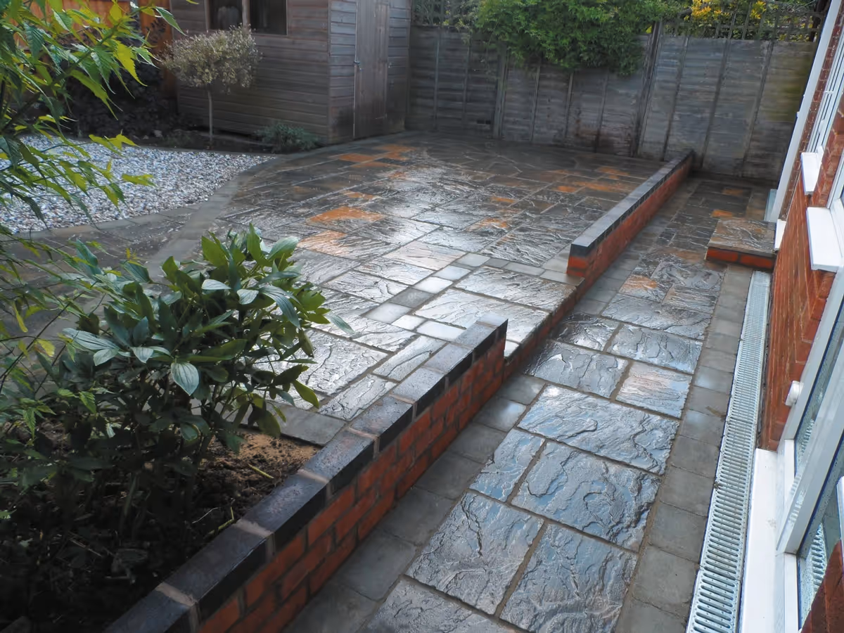 Winter stone concrete patio with matching circle, patio edged with large charcoal rumbled block, red walling finished with blue engineering brick and new lawn - angle 3