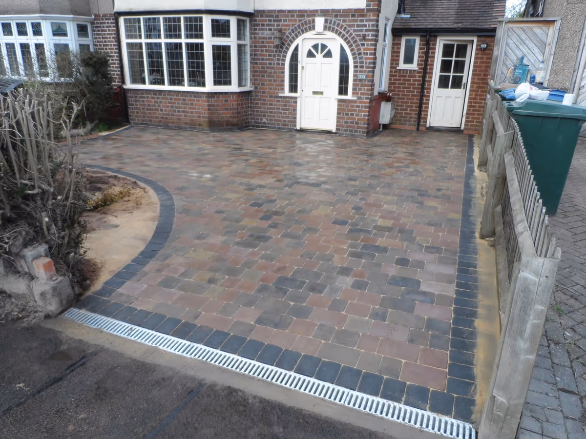 Bracken Rumbled paving set driveway with a single charcoal border, radius planter areas and an ACO channel drainage system.