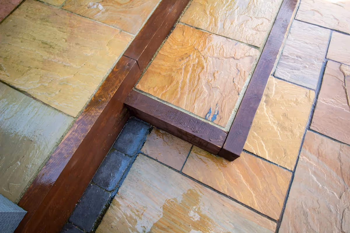 Mixed size camal natural stone patio with large charcoal rumbled block, raised sleeper area and artificial grass lawn.  - angle 3