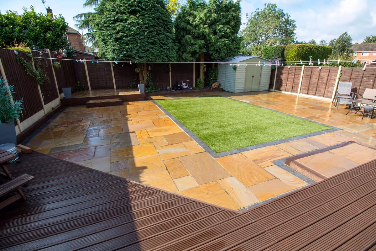 Mixed size camal natural stone patio with large charcoal rumbled block, raised sleeper area and artificial grass lawn.  - angle 5