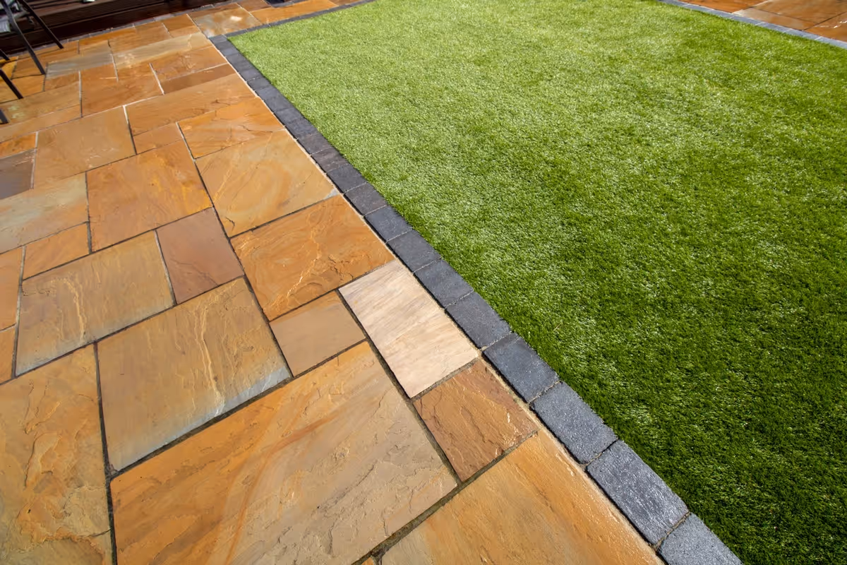 Mixed size camal natural stone patio with large charcoal rumbled block, raised sleeper area and artificial grass lawn.  - angle 7