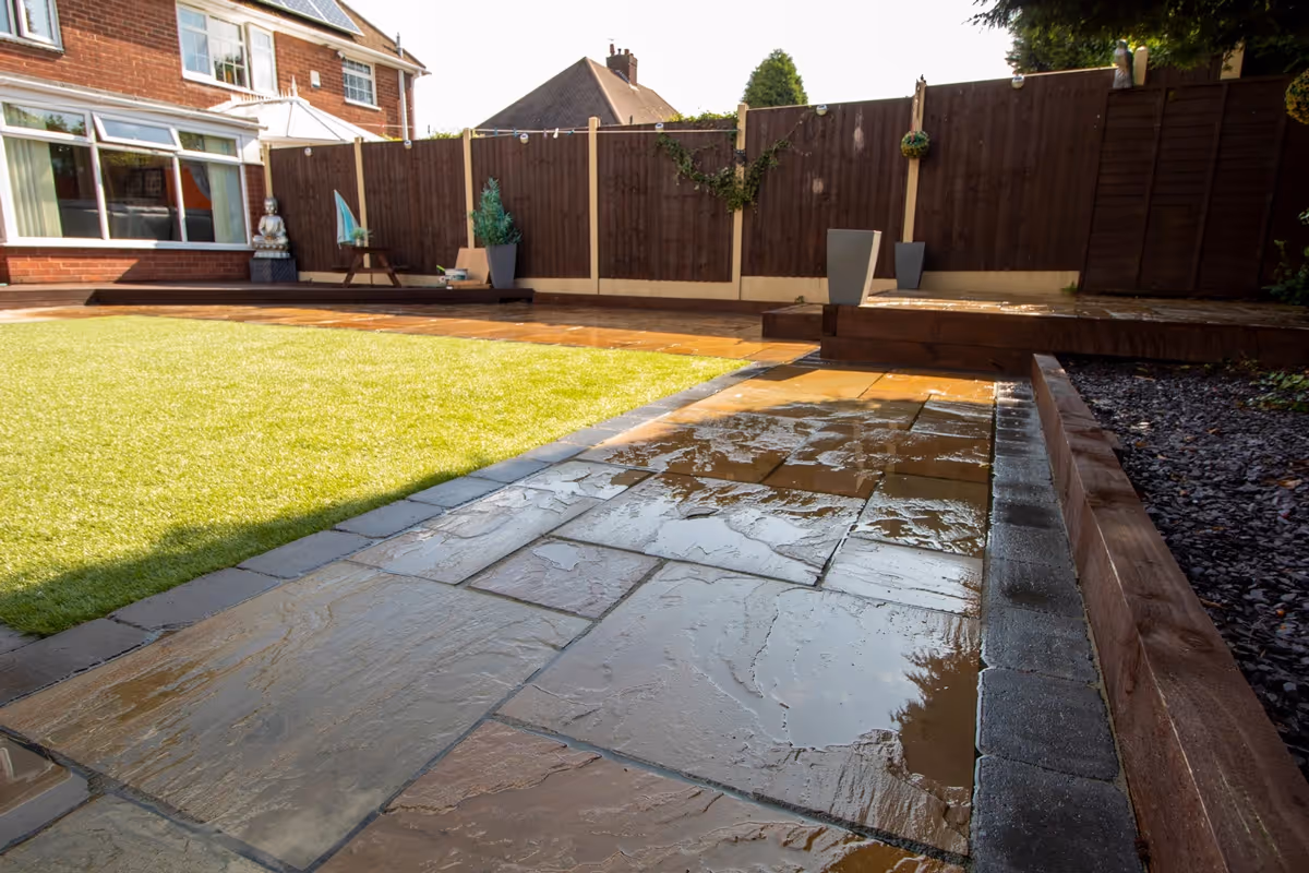 Mixed size camal natural stone patio with large charcoal rumbled block, raised sleeper area and artificial grass lawn.  - angle 2