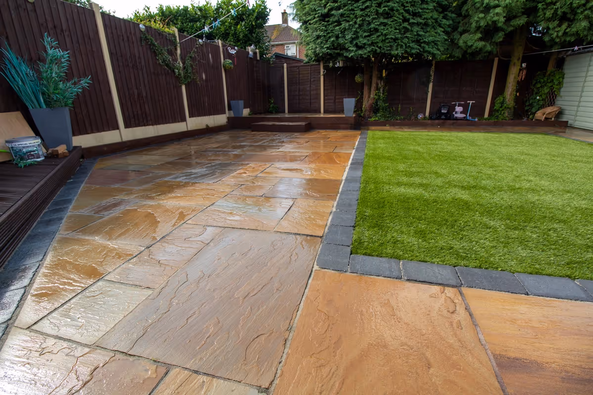 Mixed size camal natural stone patio with large charcoal rumbled block, raised sleeper area and artificial grass lawn.  - angle 1