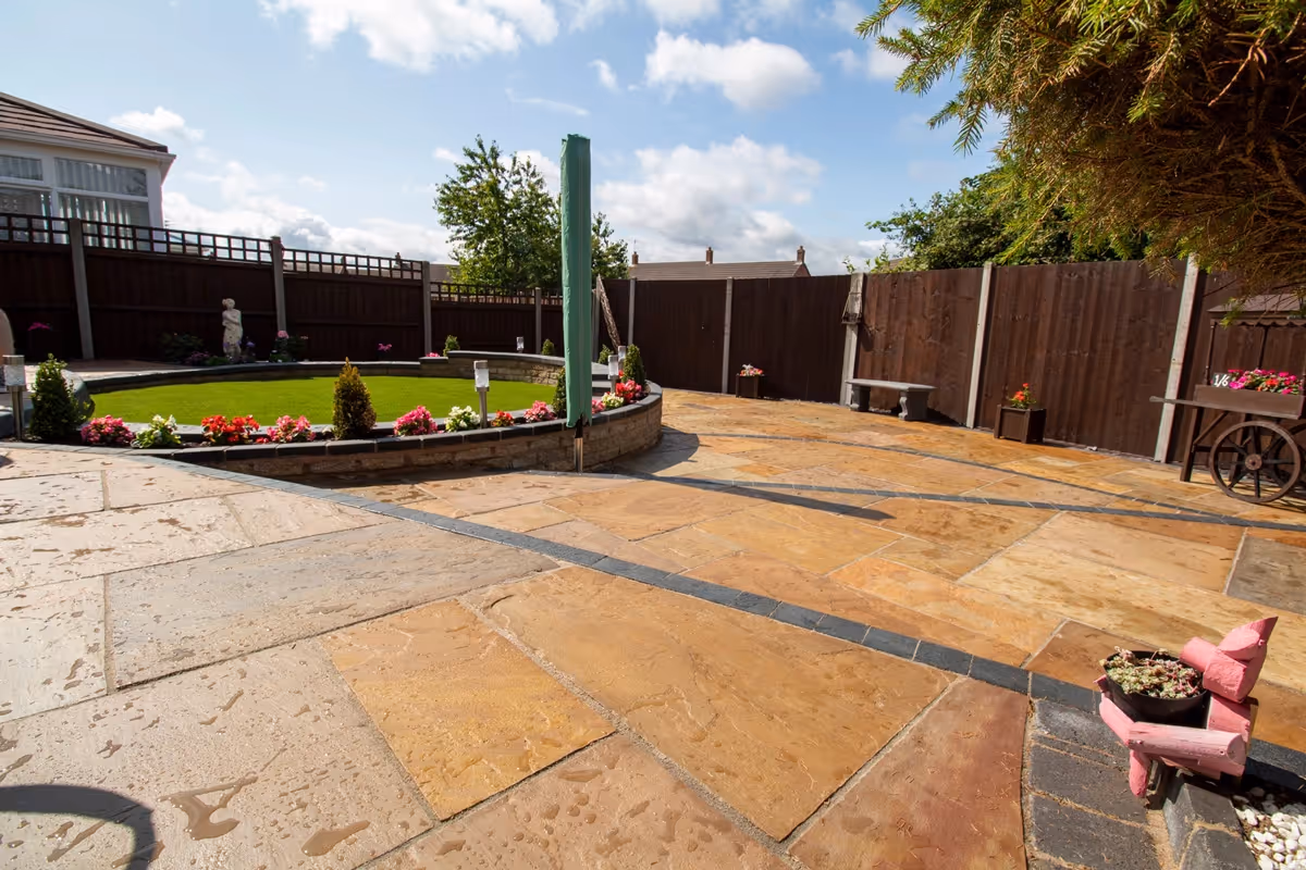 Three level camal natural stone patio with large charcoal rumbled block, steps built from charcoal kerbs, mellow York abbey walling containing a bedding area and artificial grass. -angle 2